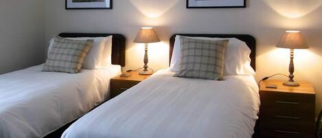 Two single beds with white linens, each with a matching pillow and a bedside table with a lamp. There are framed pictures on the wall above the beds.