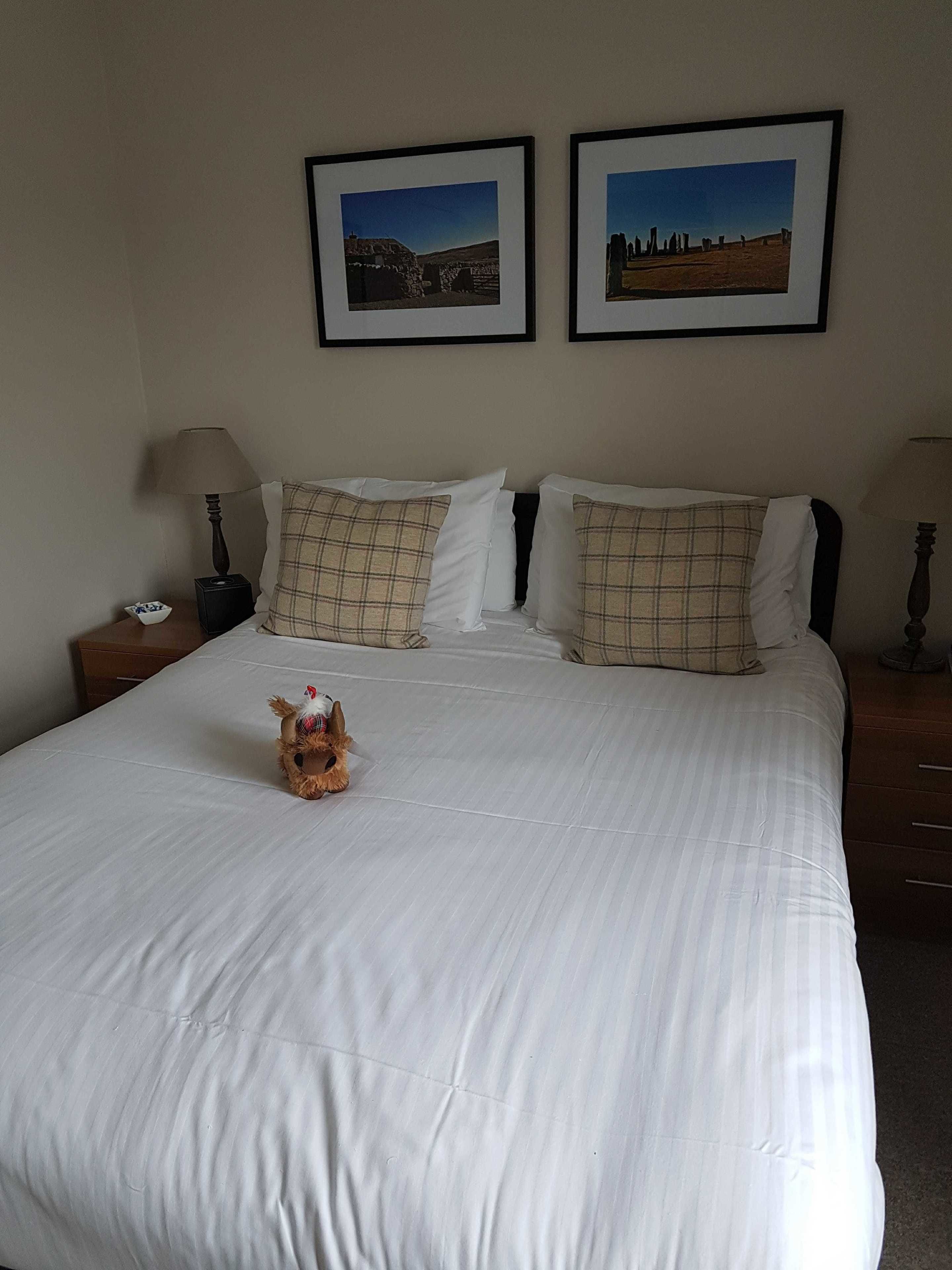 Quarto de hotel com uma cama grande, duas almofadas, uma estatueta de cachorro e duas molduras na parede.