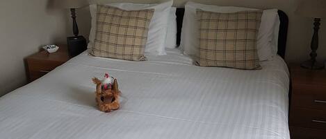 Quarto de hotel com uma cama grande, duas almofadas, uma estatueta de cachorro e duas molduras na parede.