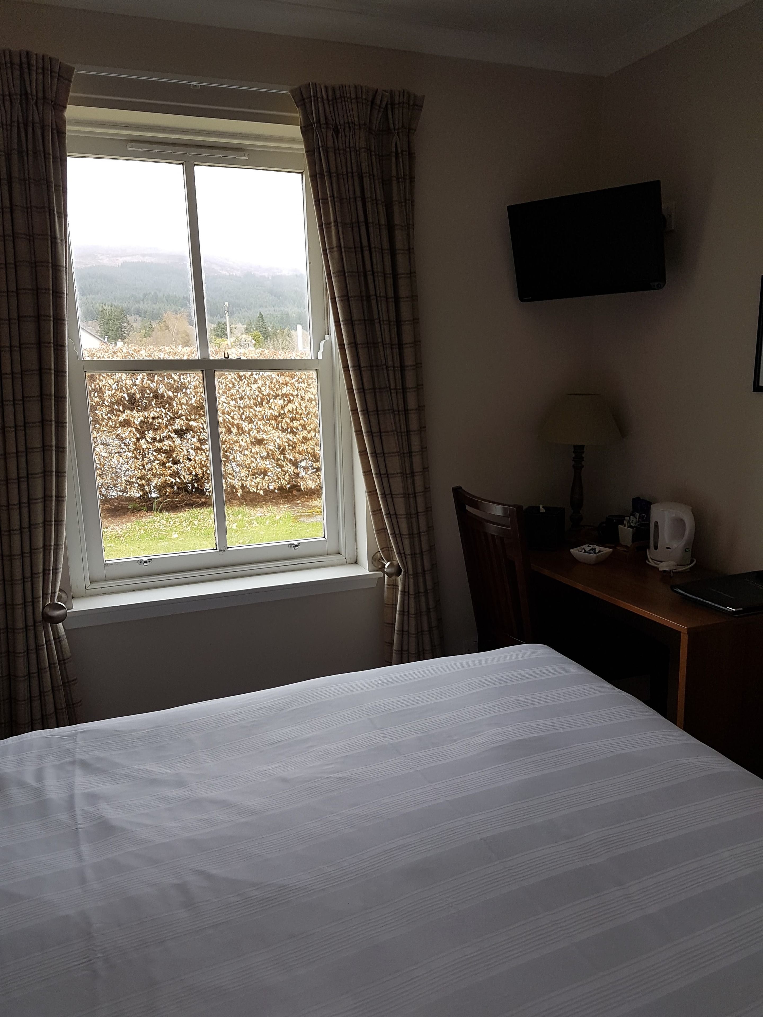 A hotel room with a bed, a desk with a lamp, a television on the wall, and a window with curtains.