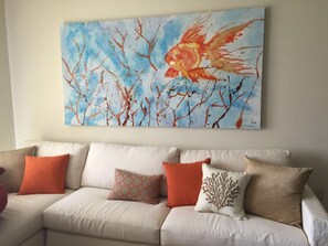 Una sala de estar con un sofá color beige, cojines naranja y blancos, y un cuadro abstracto grande en la pared.