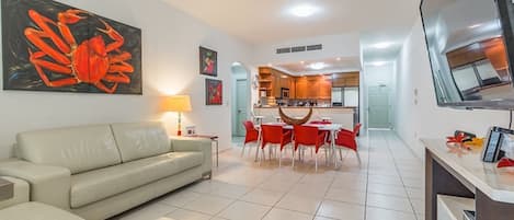 A modern living room with a sofa, dining area with red chairs, a kitchen with wooden cabinets, and a TV mounted on the wall.