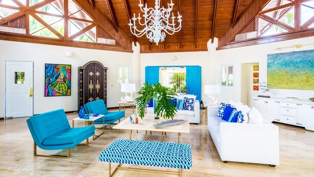 A spacious living area with a high ceiling, wooden beams, and a chandelier. There are blue and white furniture pieces, a large window with blue shutters, and artwork on the walls. - Unlimited Luxury Laureanne's Villa (La Romana)