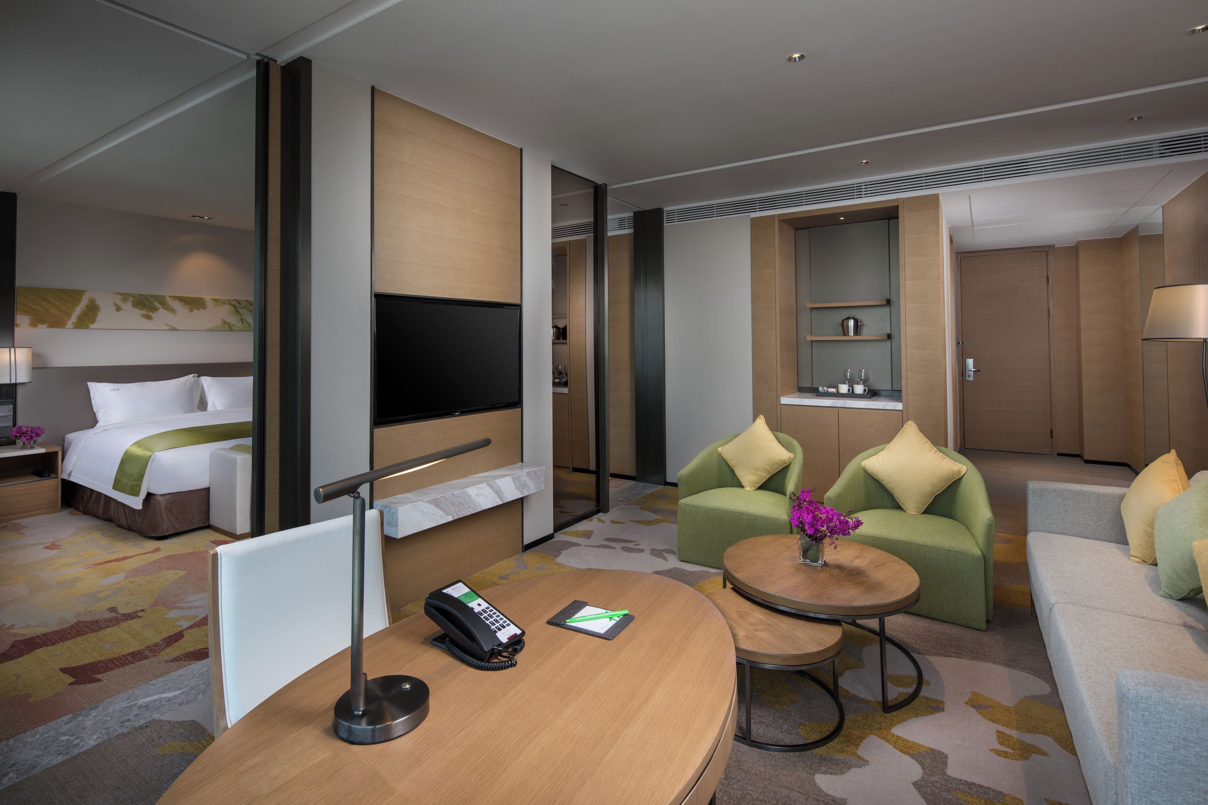 Habitación de hotel con cama, televisor, escritorio con teléfono, sofá verde, mesa redonda con un jarrón de flores y una zona de kitchenette.