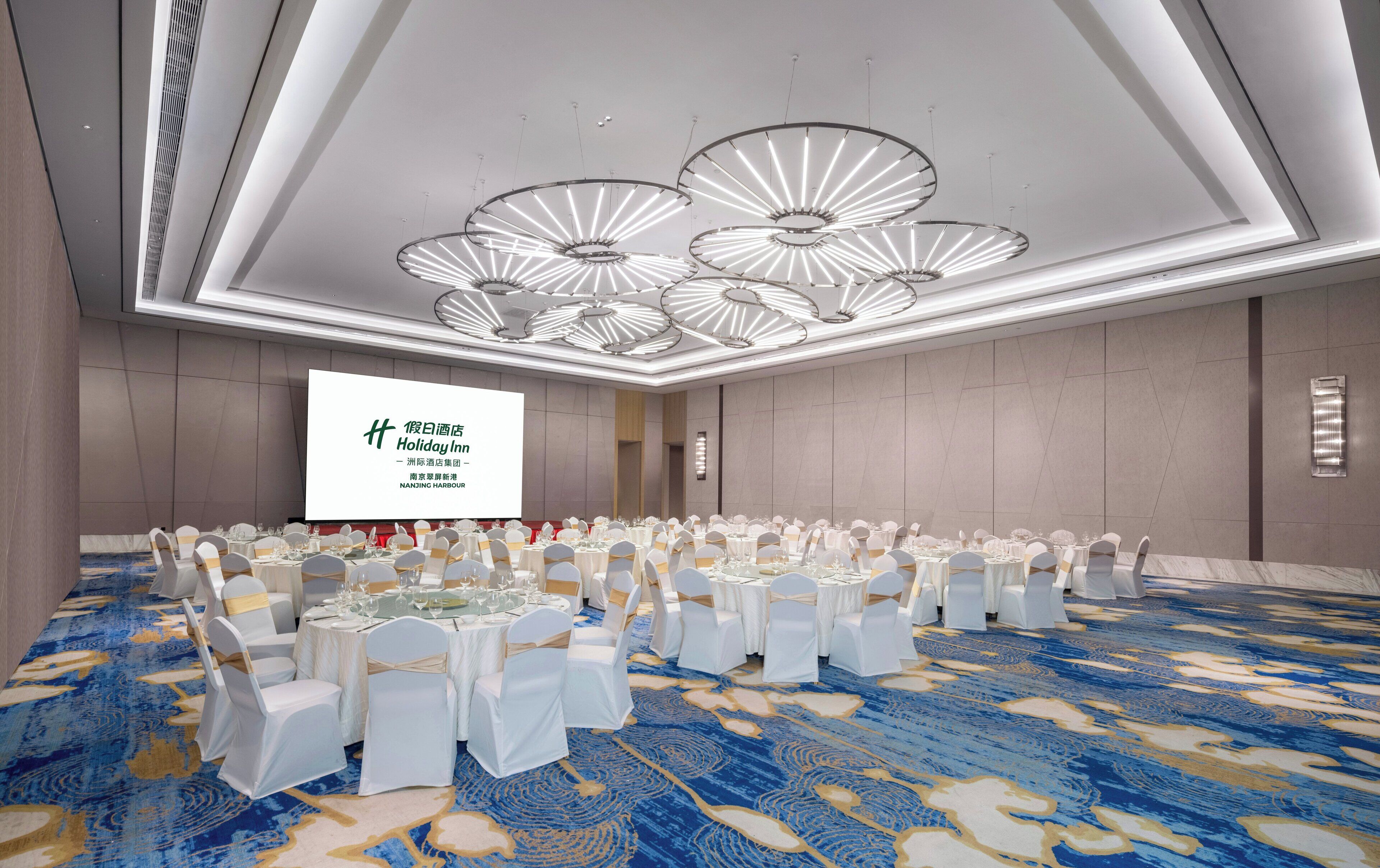 A banquet hall with round tables, white chairs, and a large screen displaying text.
