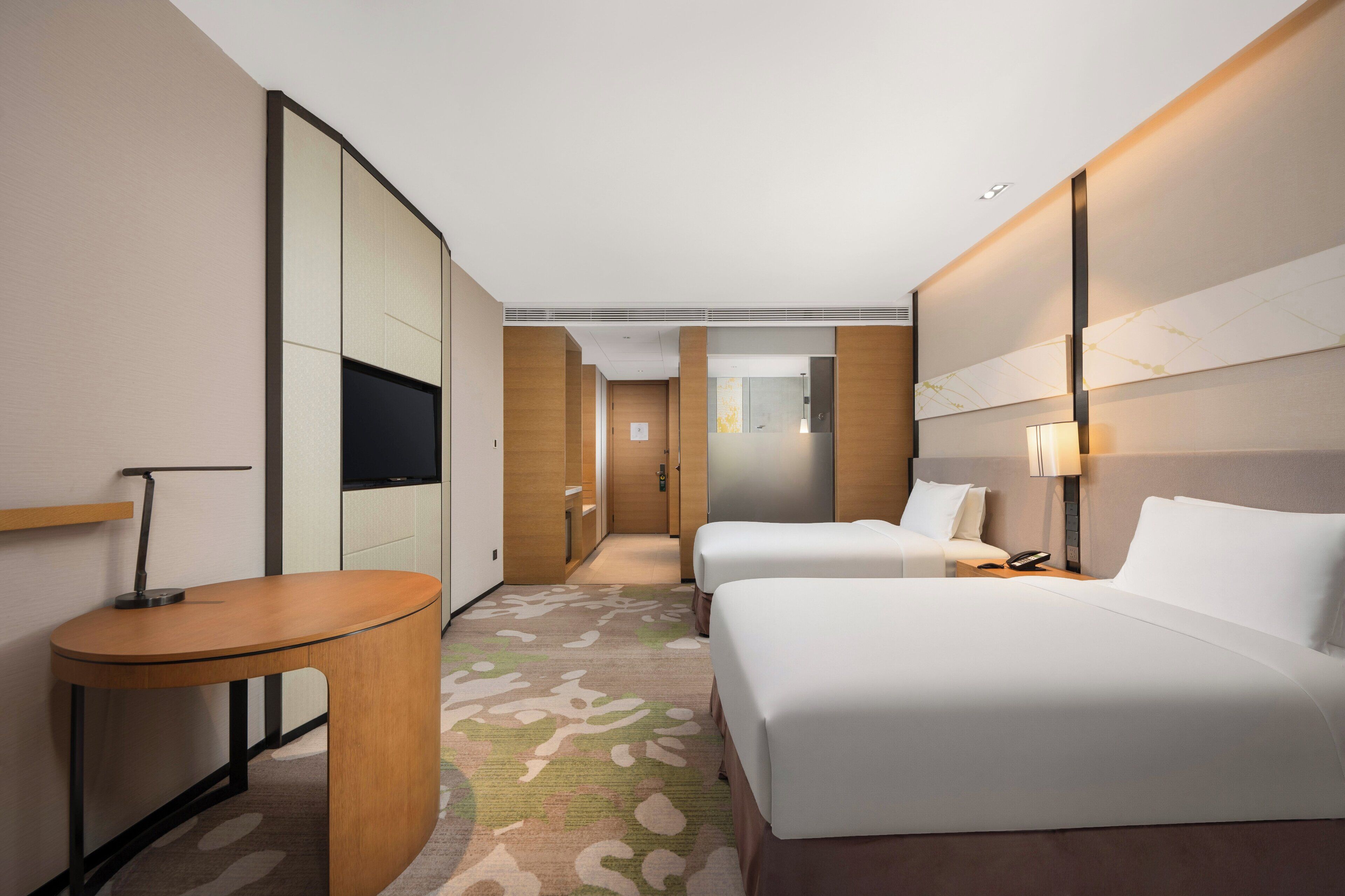 A hotel room with two beds, a desk, a TV, and a bathroom visible through a door.