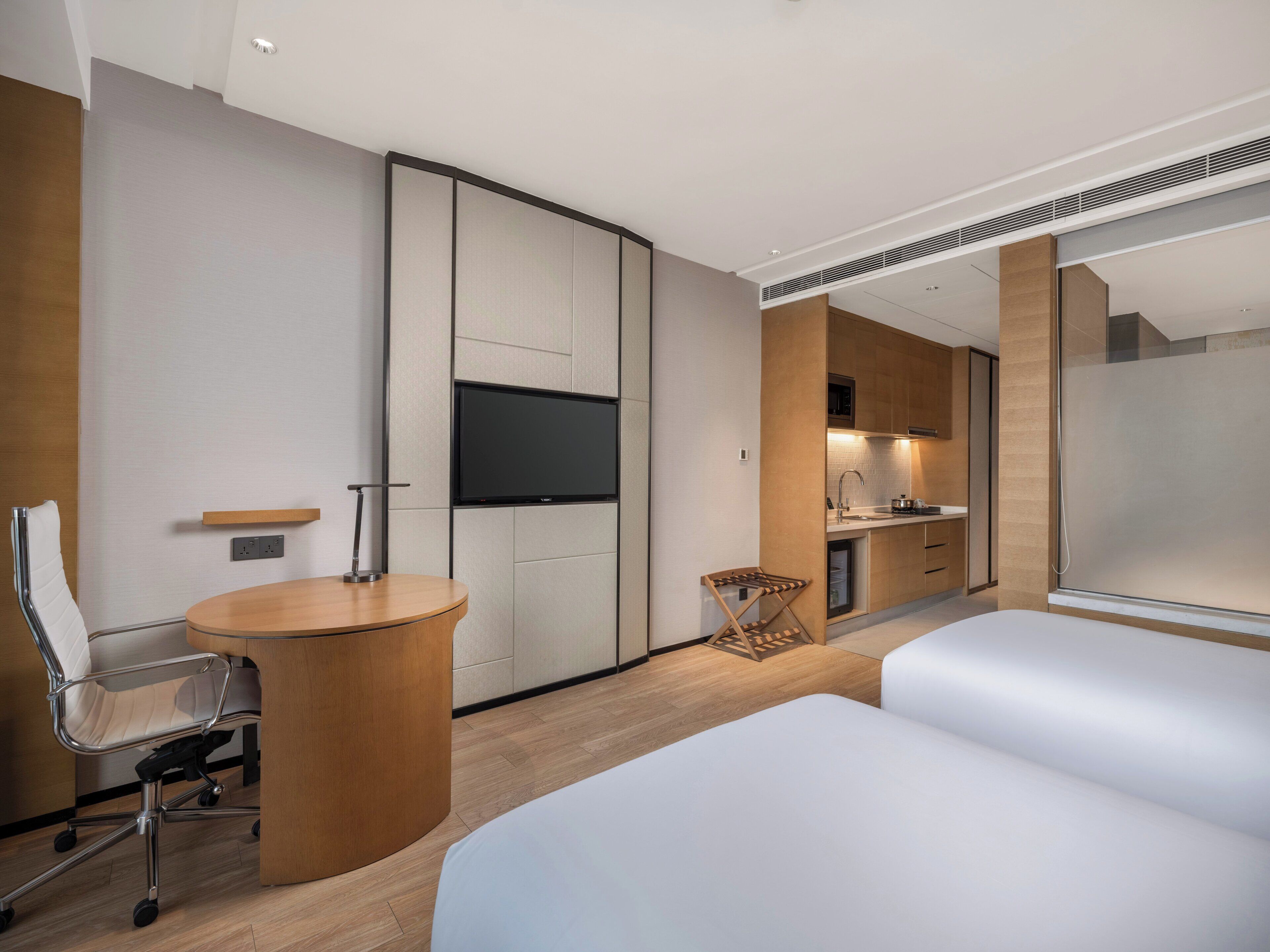 A modern hotel room with a bed, a desk, a chair, a TV, a kitchen area, and a bathroom.