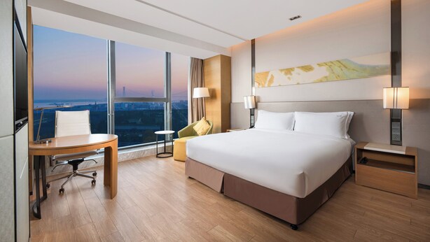 Habitación de hotel moderna con una cama grande, un escritorio, una silla y vistas de la ciudad a través de amplias ventanas.