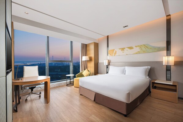 A modern hotel room with a large bed, a desk, a chair, and a view of the cityscape through large windows. - Holiday Inn Nanjing Harbour by IHG (Nanjing)