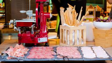 A food preparation area with a meat slicer, various meats, breadsticks, and a bowl of grapes.