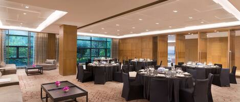 A spacious banquet hall with round tables set for dining, a sofa area, and large windows offering a view of greenery.
