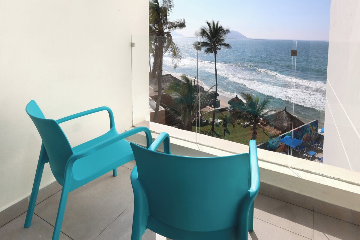 A balcony with turquoise chairs overlooking a beach with palm trees and ocean waves.