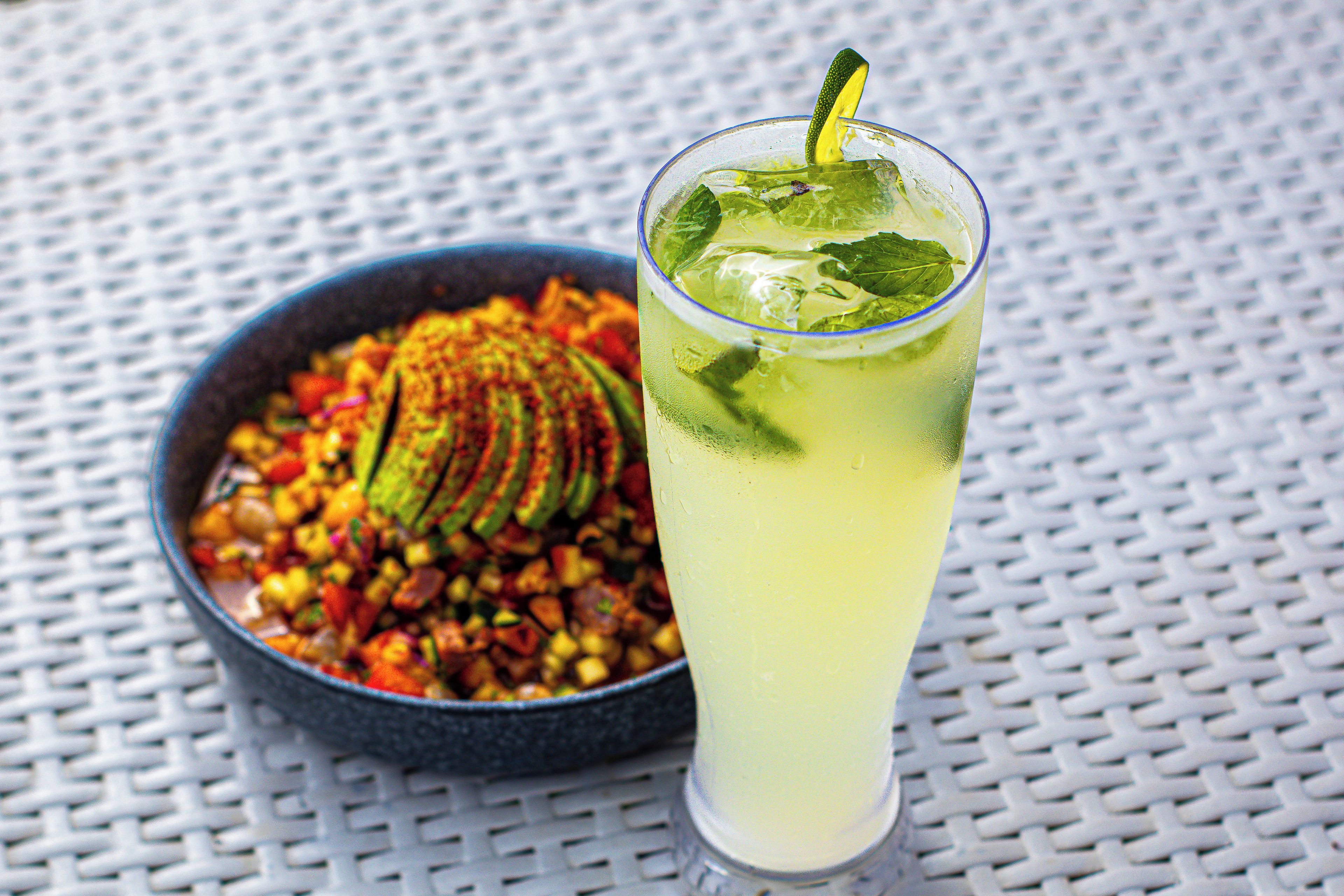 A glass of a yellow beverage with a lime slice and mint, and a bowl of colorful food next to it.