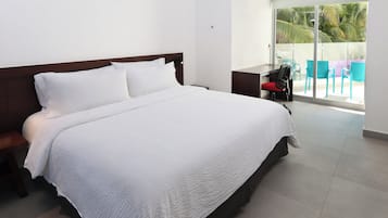 Una cama bien tendida con una colcha blanca en una habitación de hotel moderna, con un ventanal y vistas a palmeras.