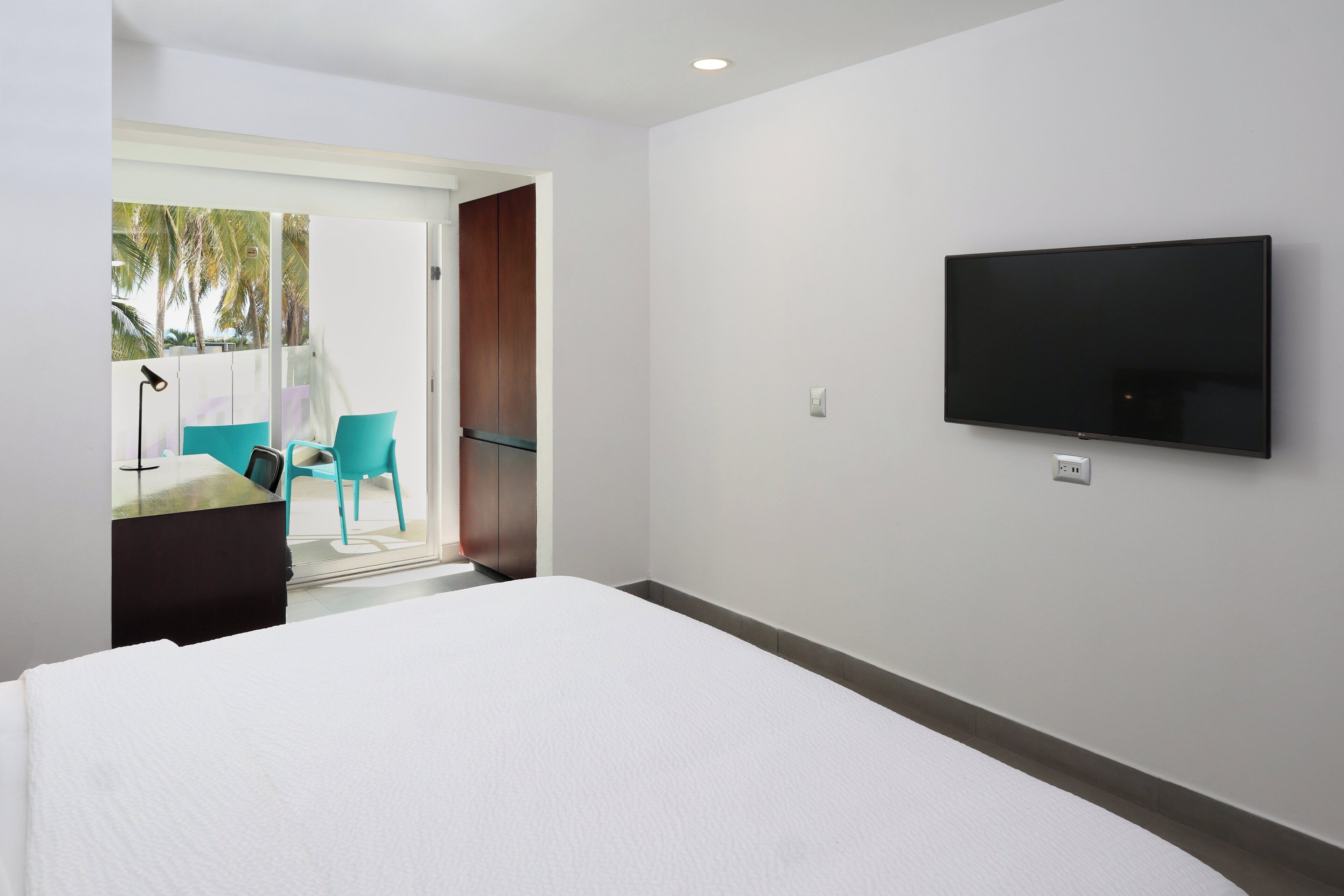 A hotel room with a bed, a TV mounted on the wall, and a door leading to a balcony with chairs and a table.