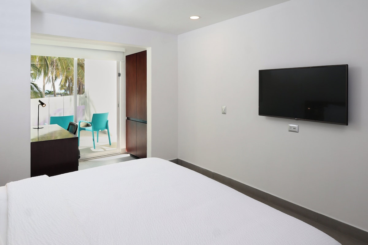 A hotel room with a bed, a TV mounted on the wall, and a door leading to a balcony with chairs and a table.
