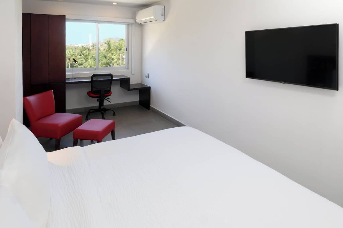 Habitación de hotel con cama, escritorio con silla, televisor y ventana con vista a palmeras.