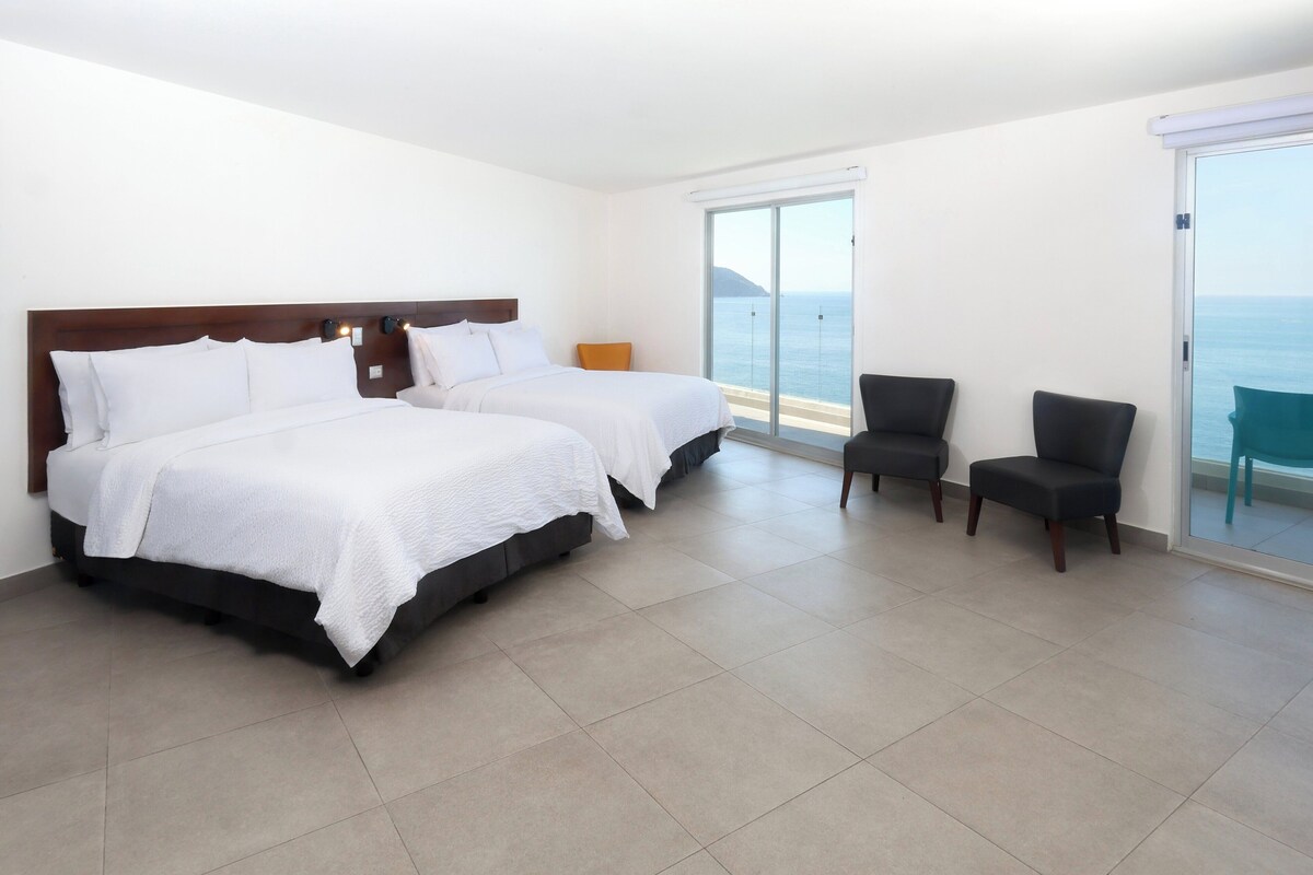 A hotel room with two beds, a seating area, and a view of the sea.