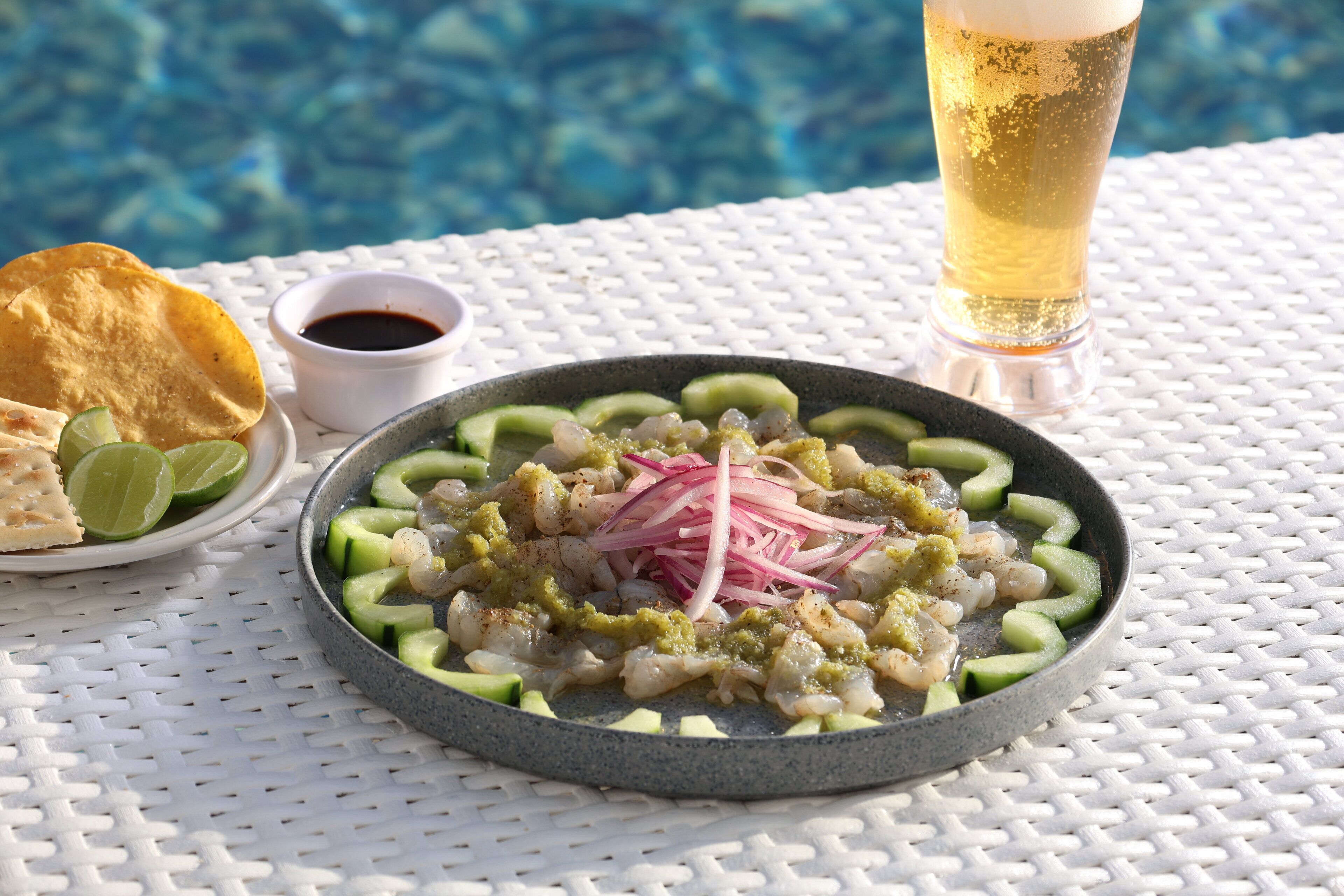 Un plat de fruits de mer agrémenté de citron vert et d’oignons, un verre de bière et un petit bol de sauce, le tout posé sur une table blanche au bord d’une piscine.