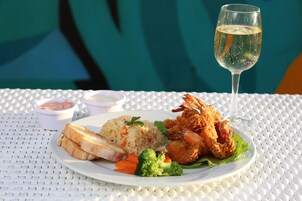 Un plato de comida con arroz, camarones fritos y pan, acompañado de una copa de vino blanco, sobre una mesa blanca.