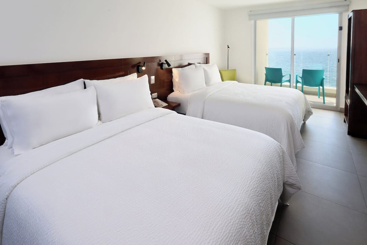 Habitación de hotel con dos camas, una mesita de noche, una silla verde y vista al mar.