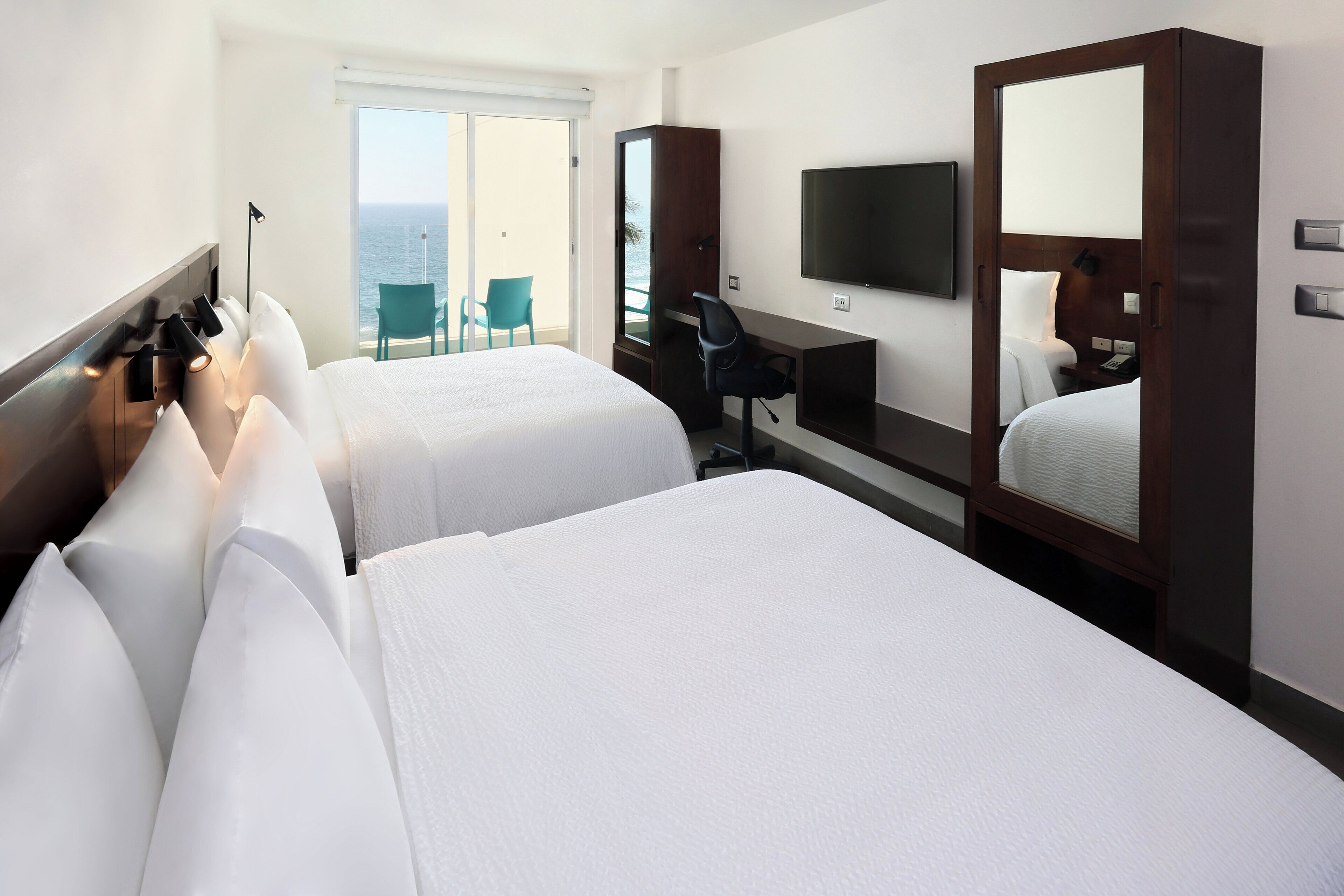 Une chambre d’hôtel avec deux lits, un bureau, une chaise, une télévision et un balcon avec vue sur la mer.