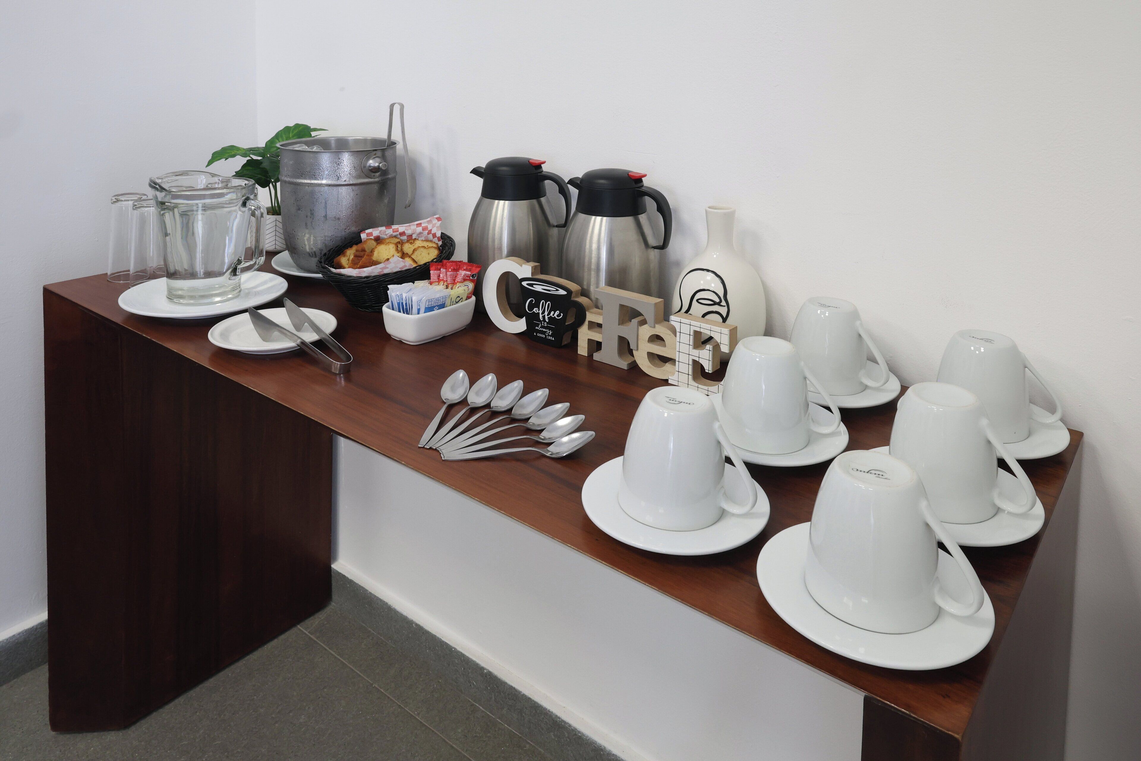 Montaje para el desayuno con cafeteras, teteras, tazas y cubiertos sobre una mesa de madera.