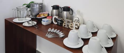 Montaje para el desayuno con cafeteras, teteras, tazas y cubiertos sobre una mesa de madera.