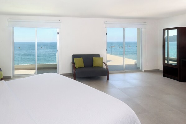 Habitación de hotel con una cama grande, una silla con cojines, un televisor y un balcón con vistas al mar.