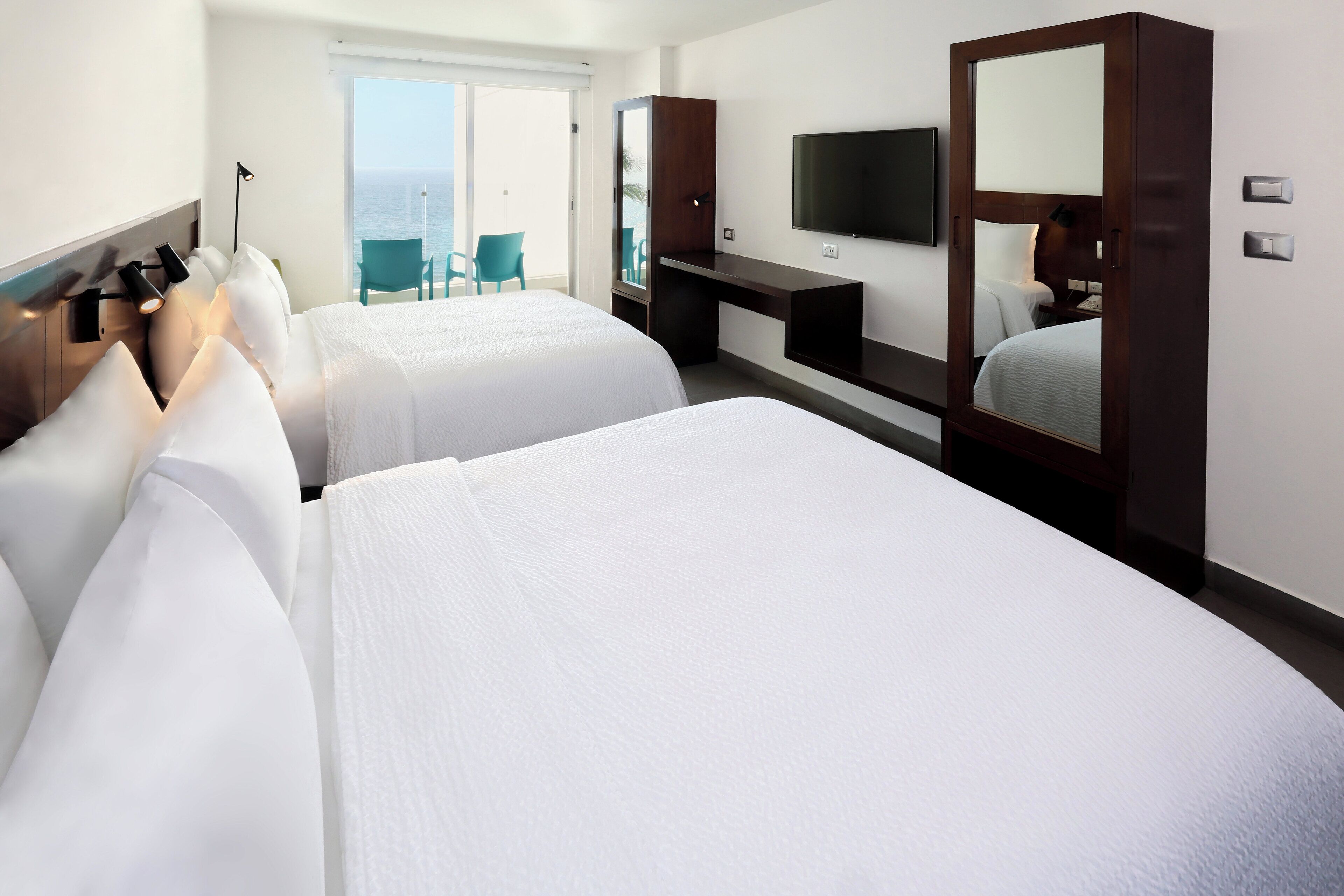 A hotel room with two beds, a TV, a desk, and a balcony with chairs.