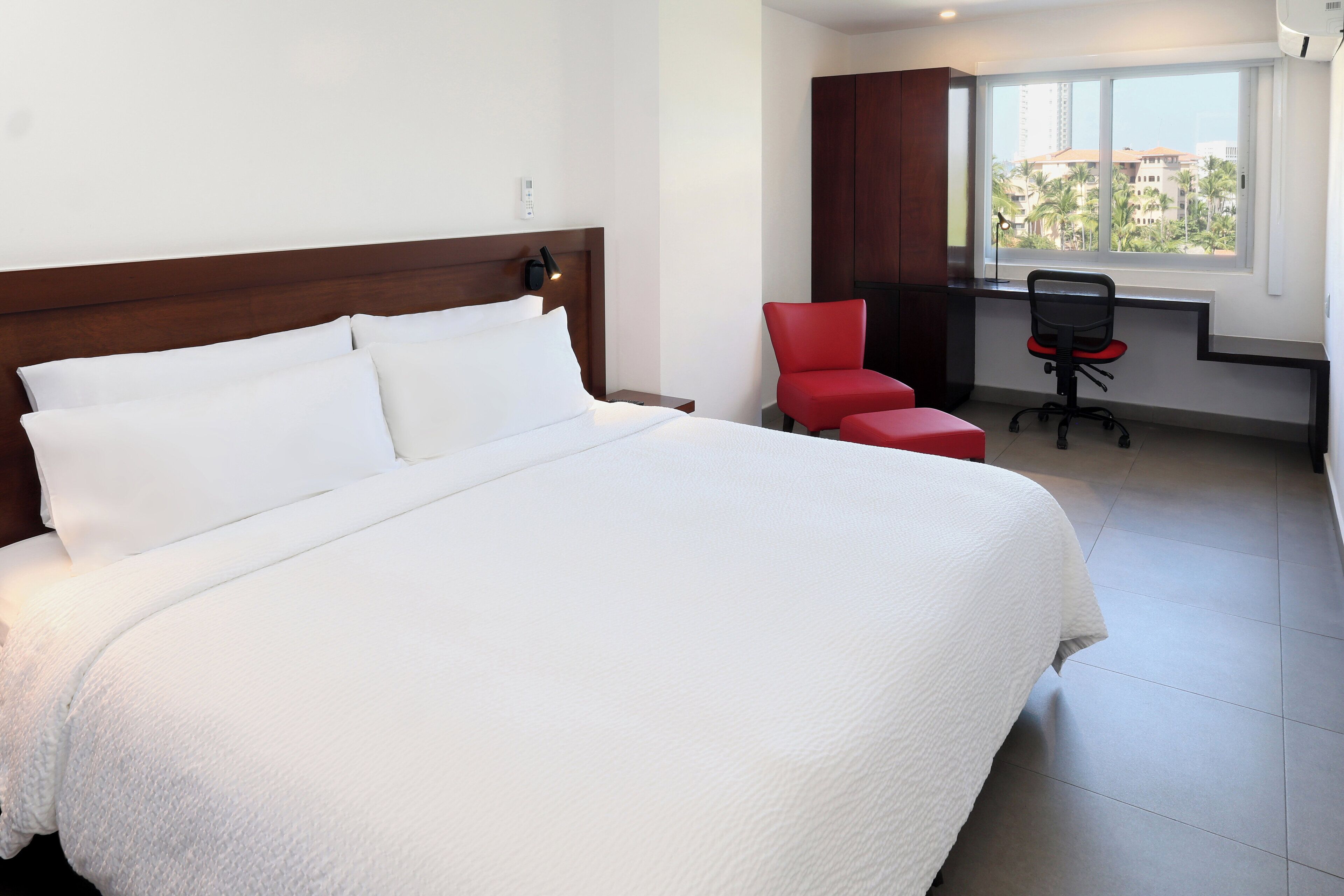 A hotel room with a large bed, a red chair, a desk, and a window with a view of buildings and palm trees.