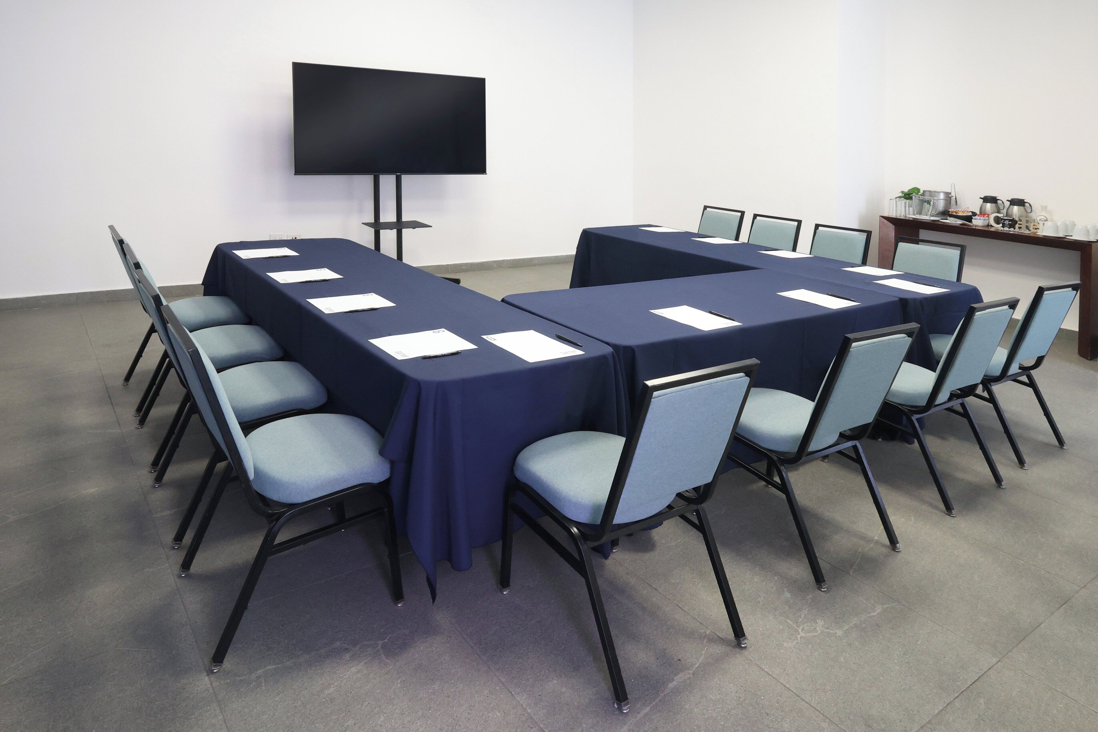 A conference room with two long tables, chairs, a flat-screen TV, and a coffee station.