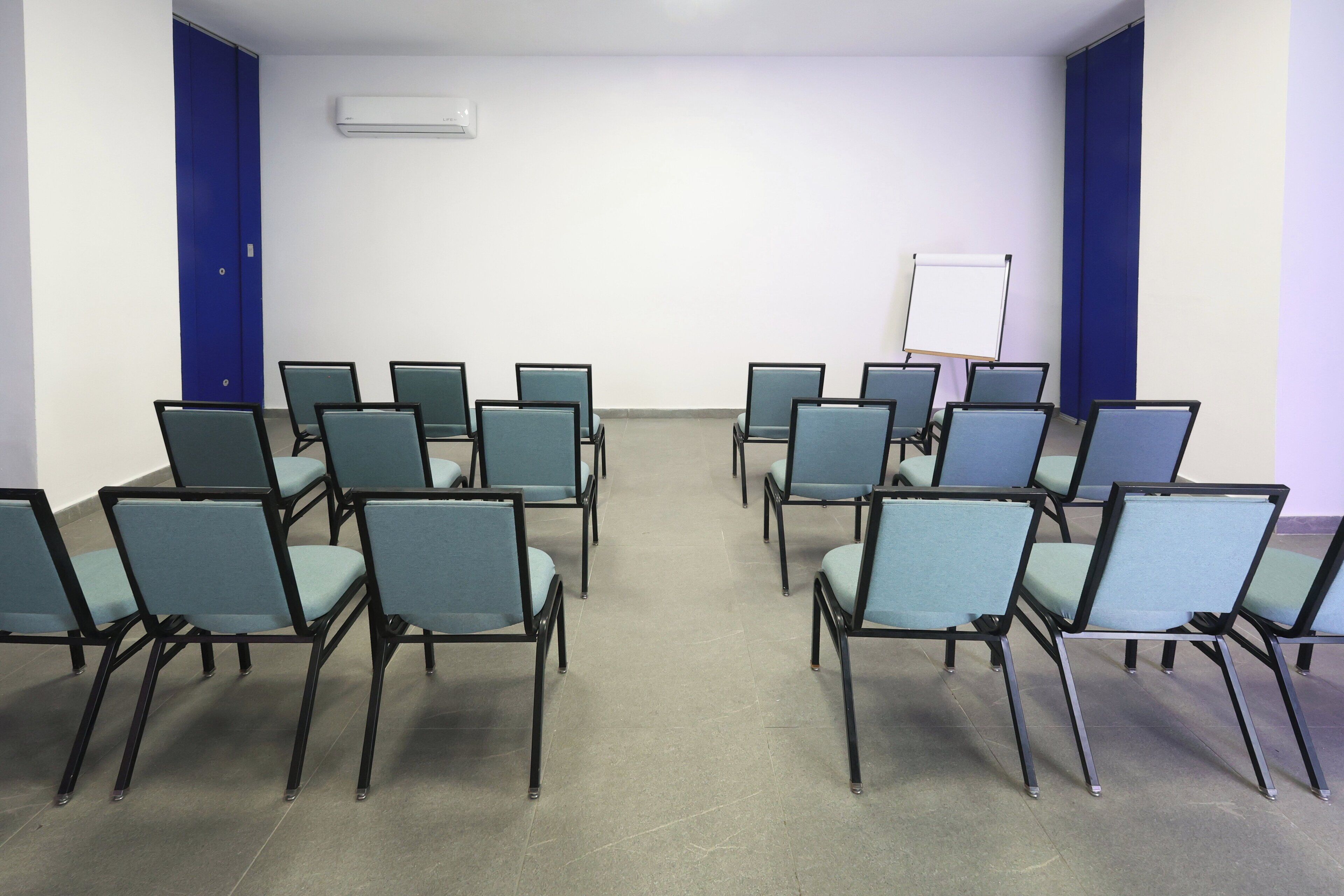 Une salle de conférence avec des rangées de chaises, un tableau blanc et des rideaux bleus.