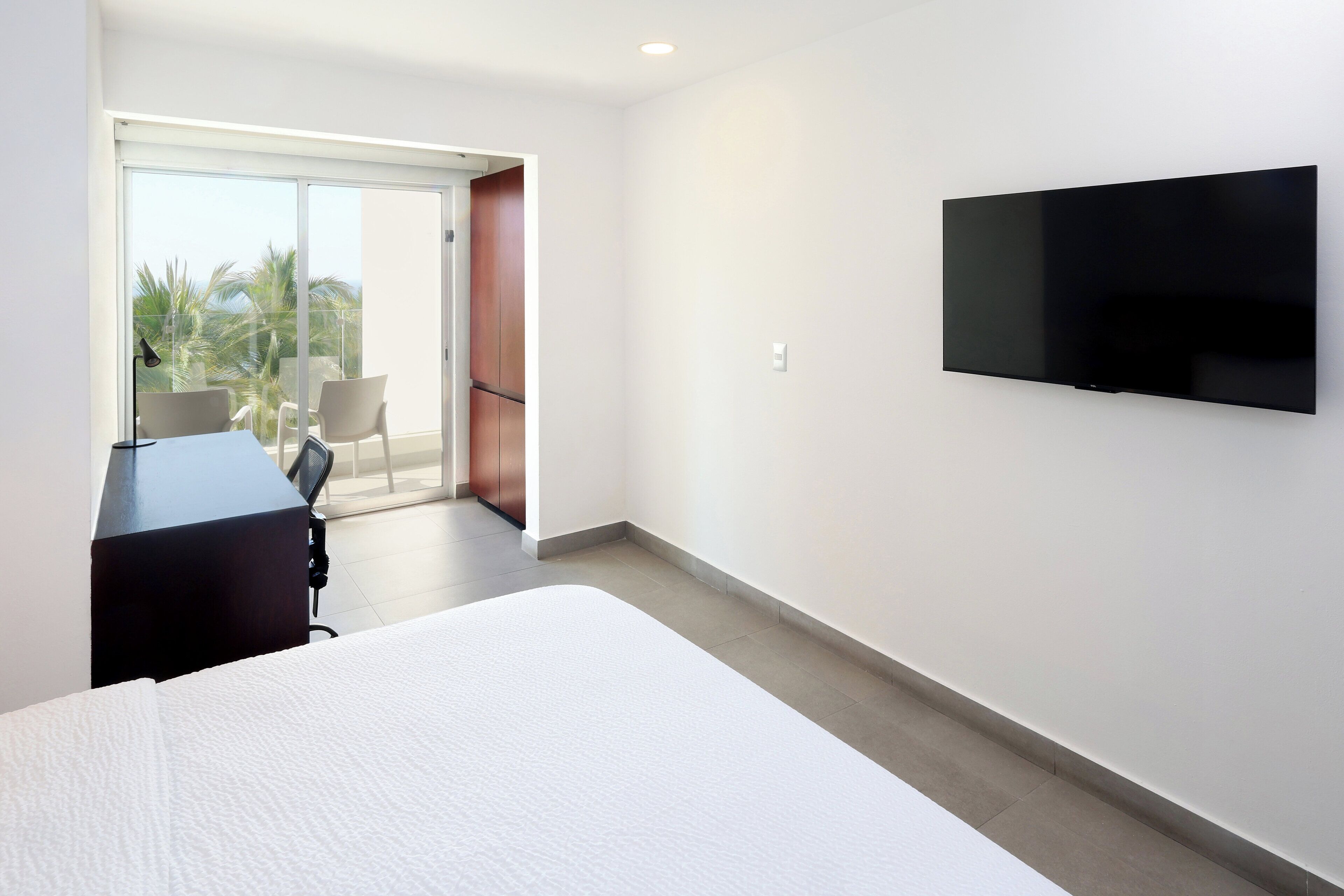 Une chambre d’hôtel moderne dotée d’un grand lit, d’un bureau avec une chaise, d’un téléviseur à écran plat fixé au mur et d’une porte-fenêtre coulissante donnant sur un balcon avec vue sur les palmiers.