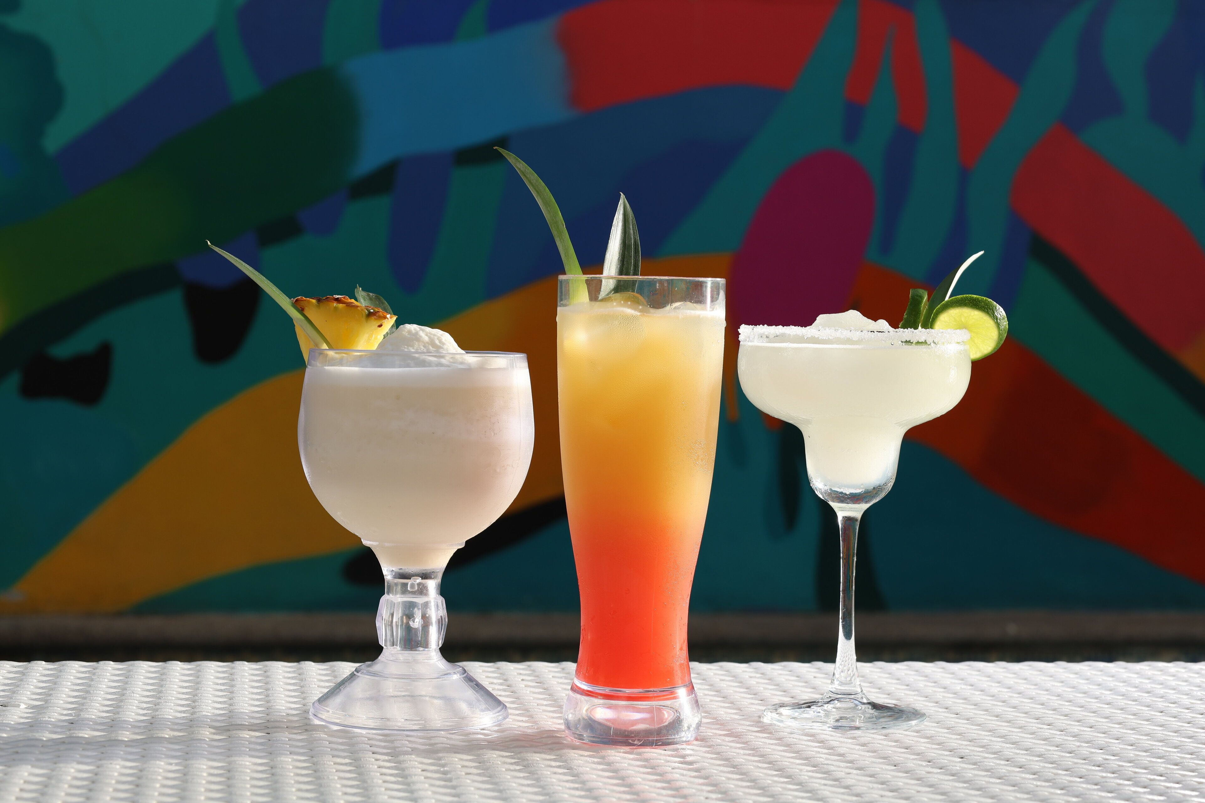 Drei Cocktails auf einem weißen Tisch mit farbigem, abstraktem Hintergrund.