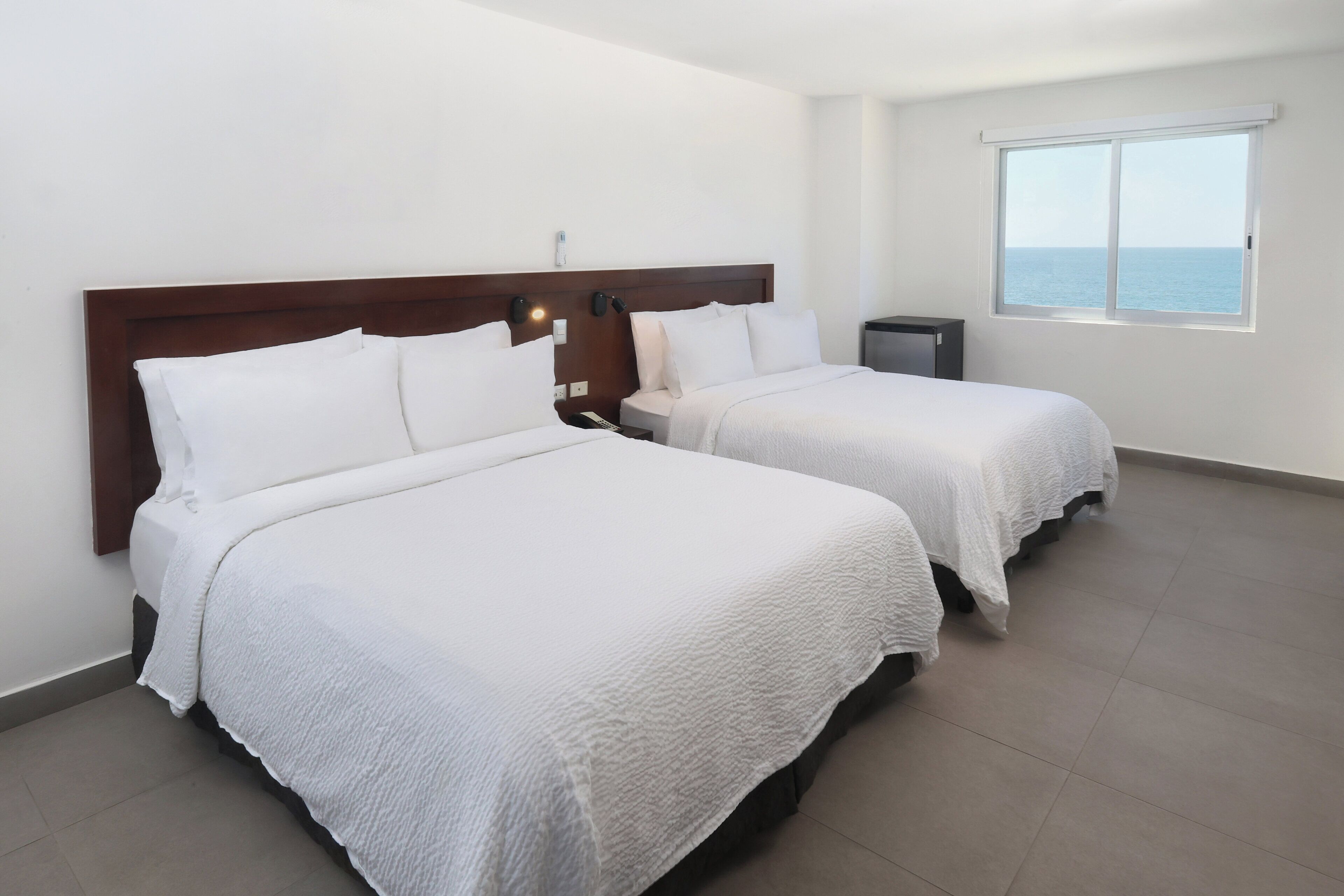 A hotel room with two beds, a nightstand, a small trash bin, and a window with a view of the sea.