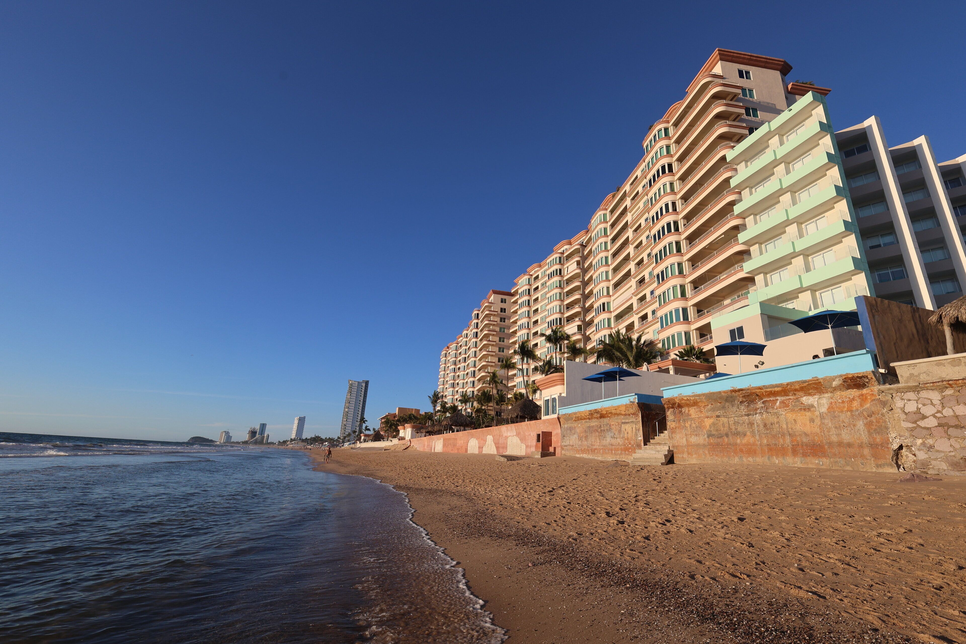 A beachfront hotel with multiple floors, a sandy beach, and a clear blue sky.