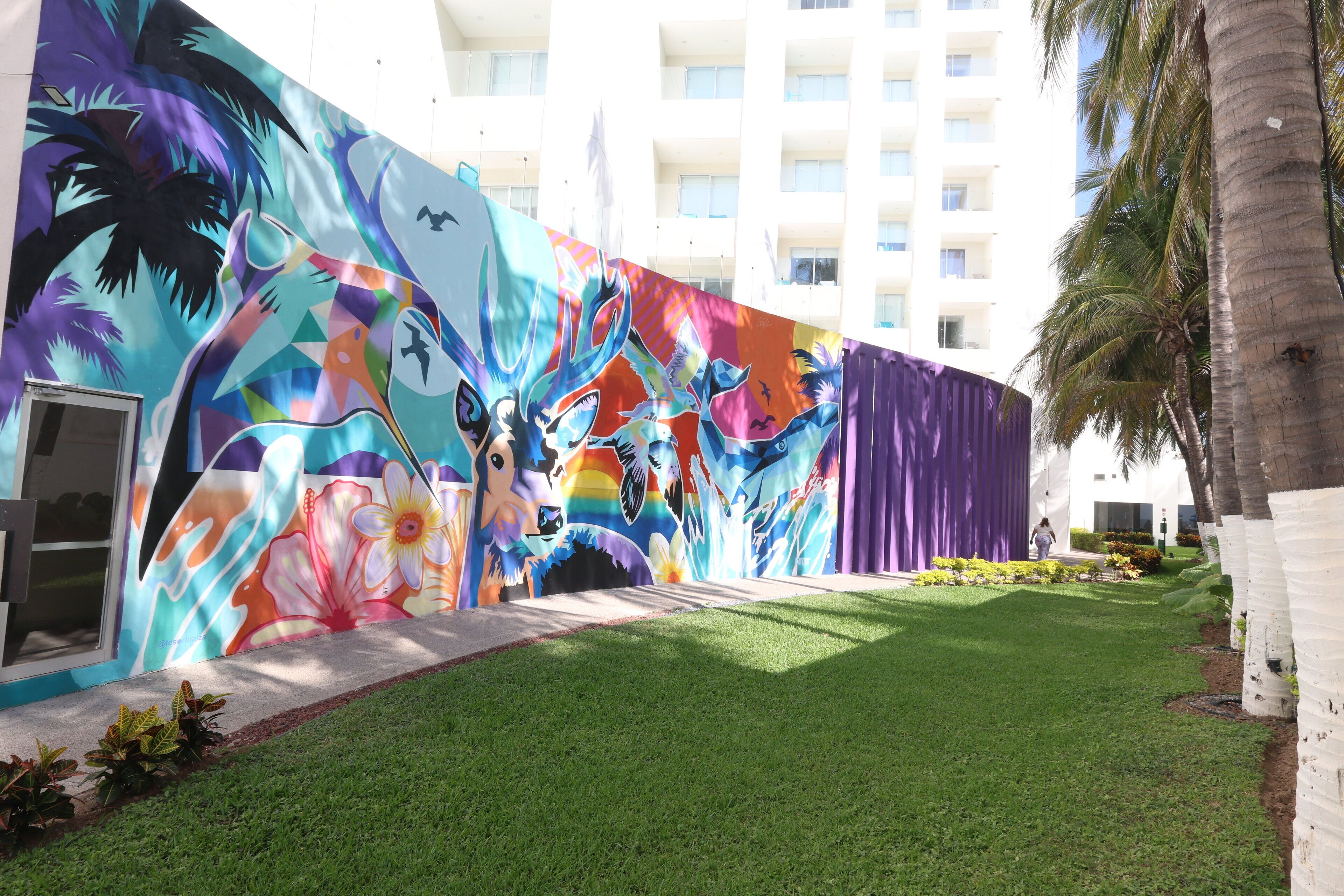 Ein Gebäude mit farbenfrohem Wandbild an der Außenwand, einem gepflegten Rasen und einer hohen Palme.