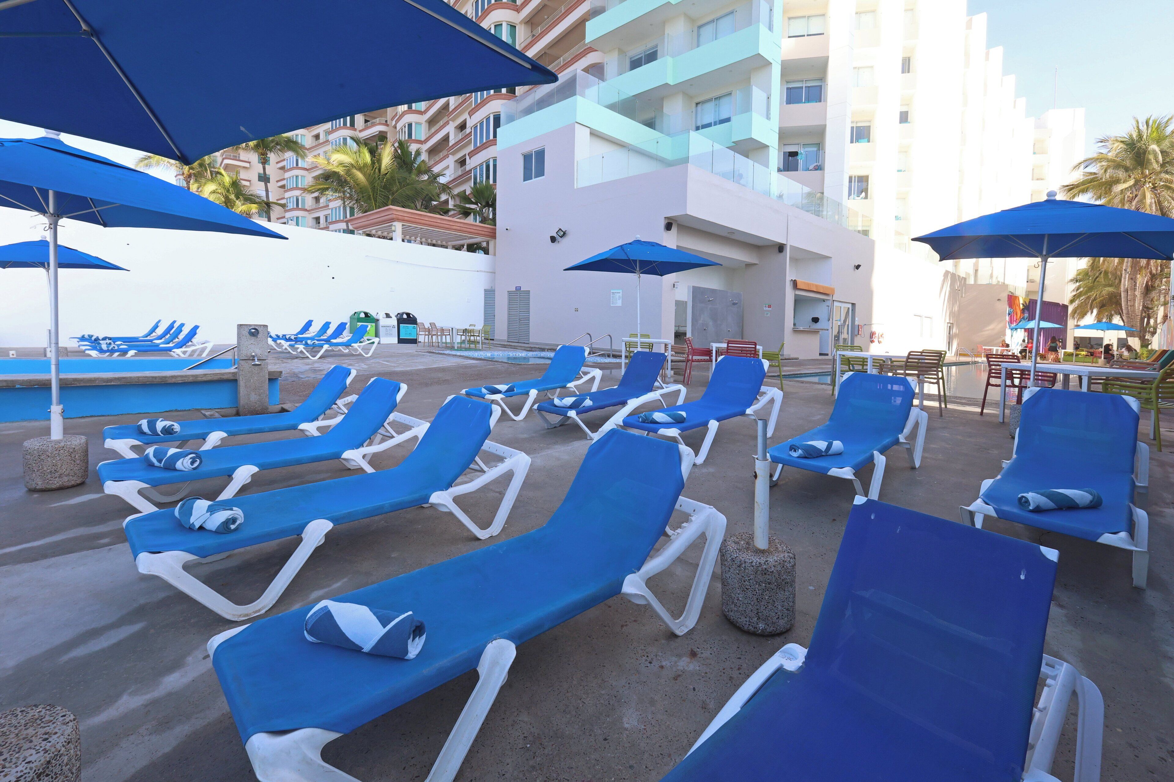 Un espace aménagé au bord de la piscine, avec des chaises longues bleues et des parasols, une piscine, et un bâtiment avec des balcons en arrière-plan.