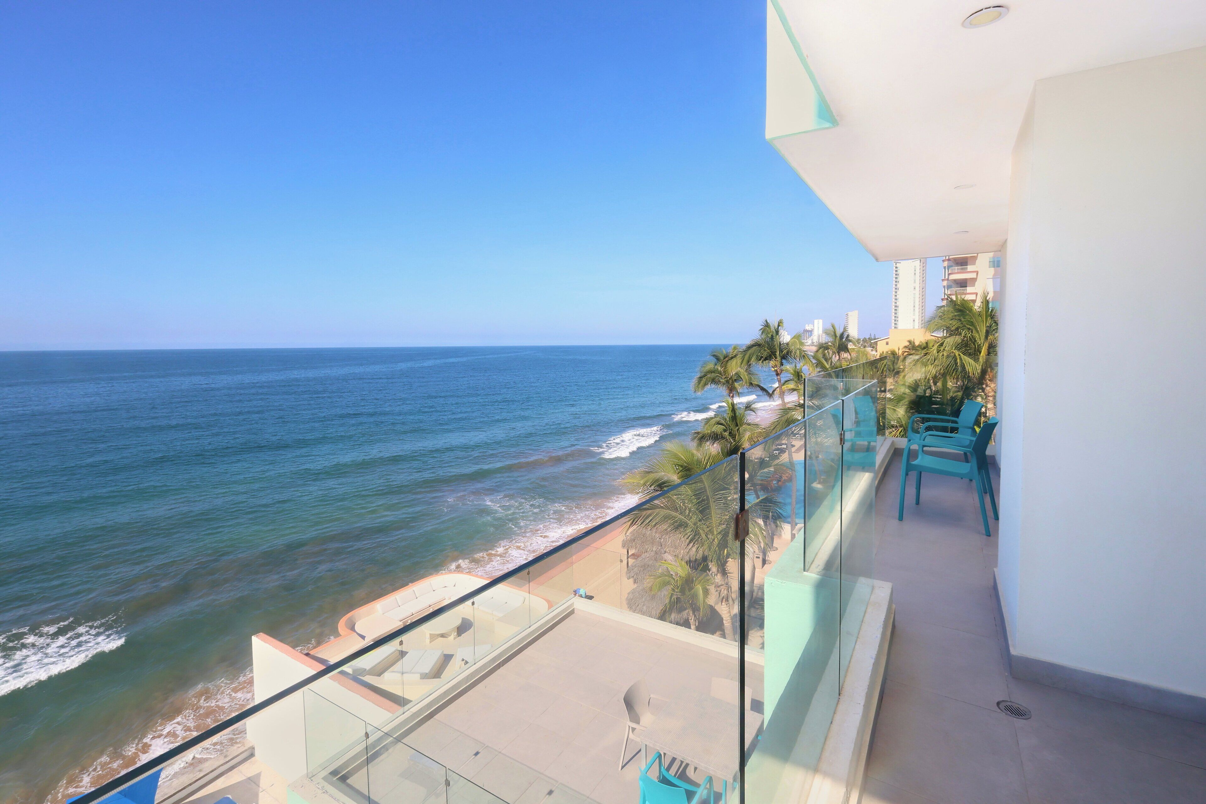 A balcony with a view of the ocean and beach, glass railings, and outdoor seating.