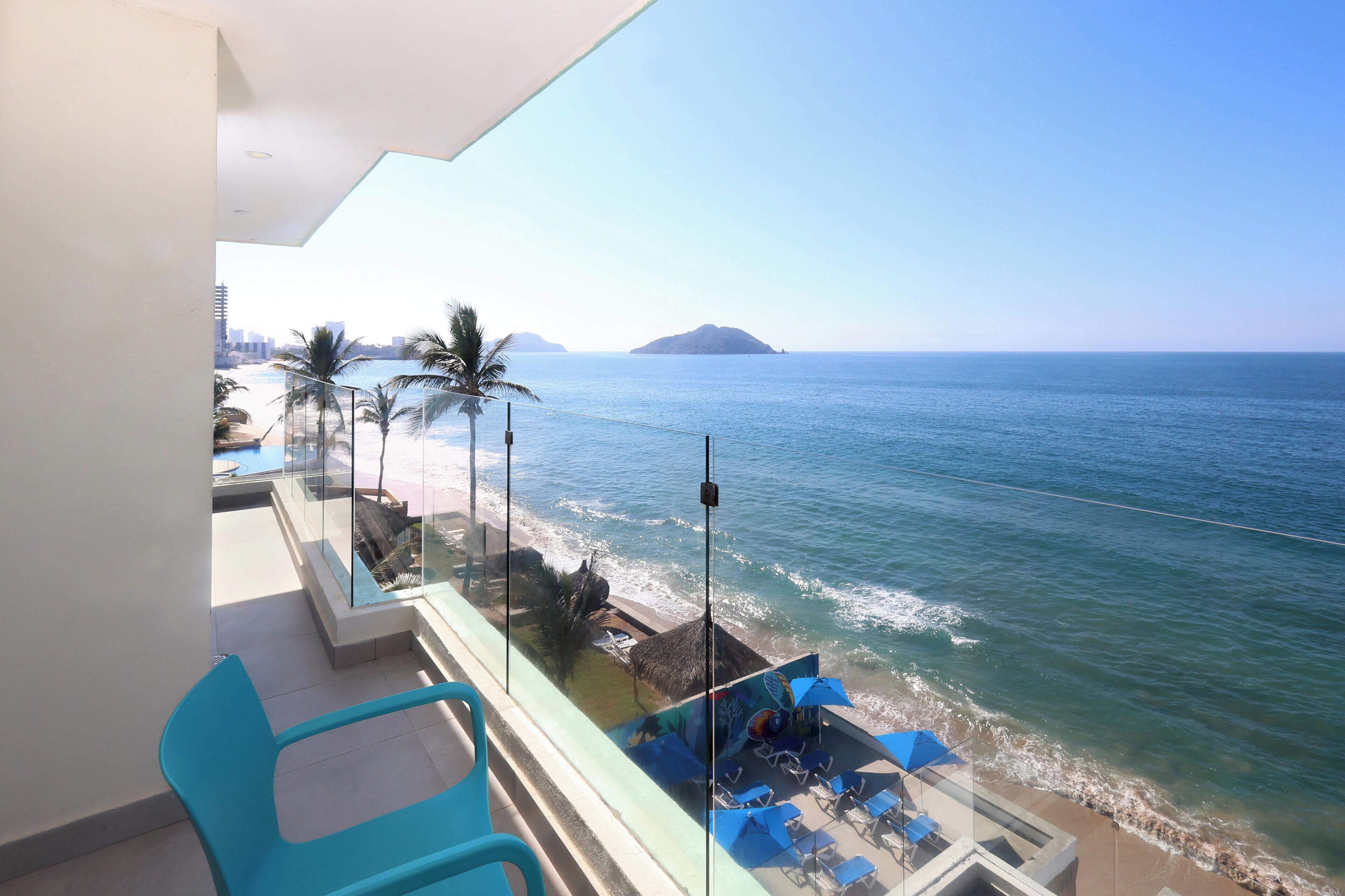 A balcony with a view of the sea, a lounge area with blue chairs and umbrellas, and a glass railing.