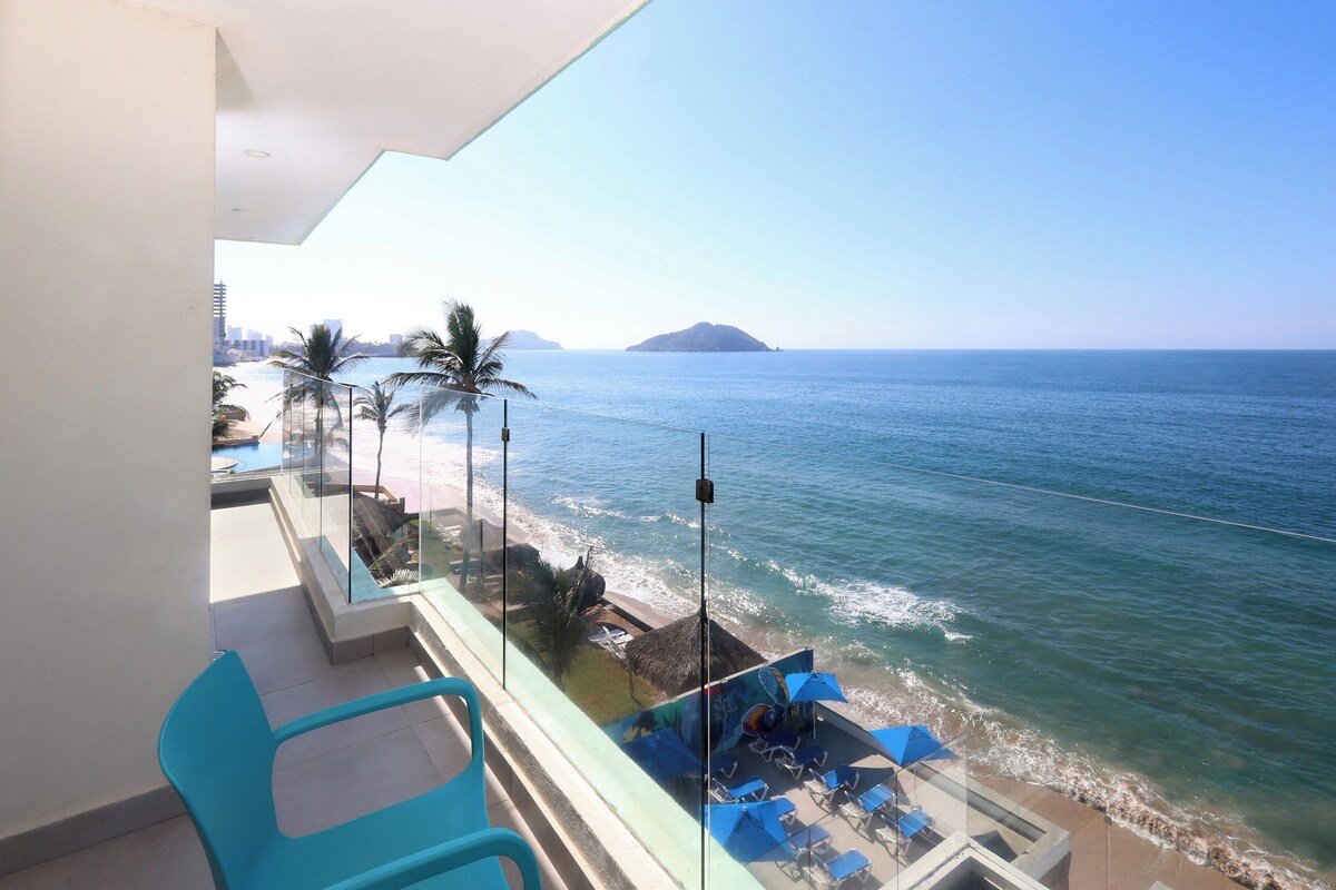 A balcony with a view of the sea, a lounge area with blue chairs and umbrellas, and a glass railing.