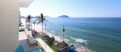 Un balcón con vistas al mar, un área de descanso con sillas y sombrillas azules, y una barandilla de vidrio.