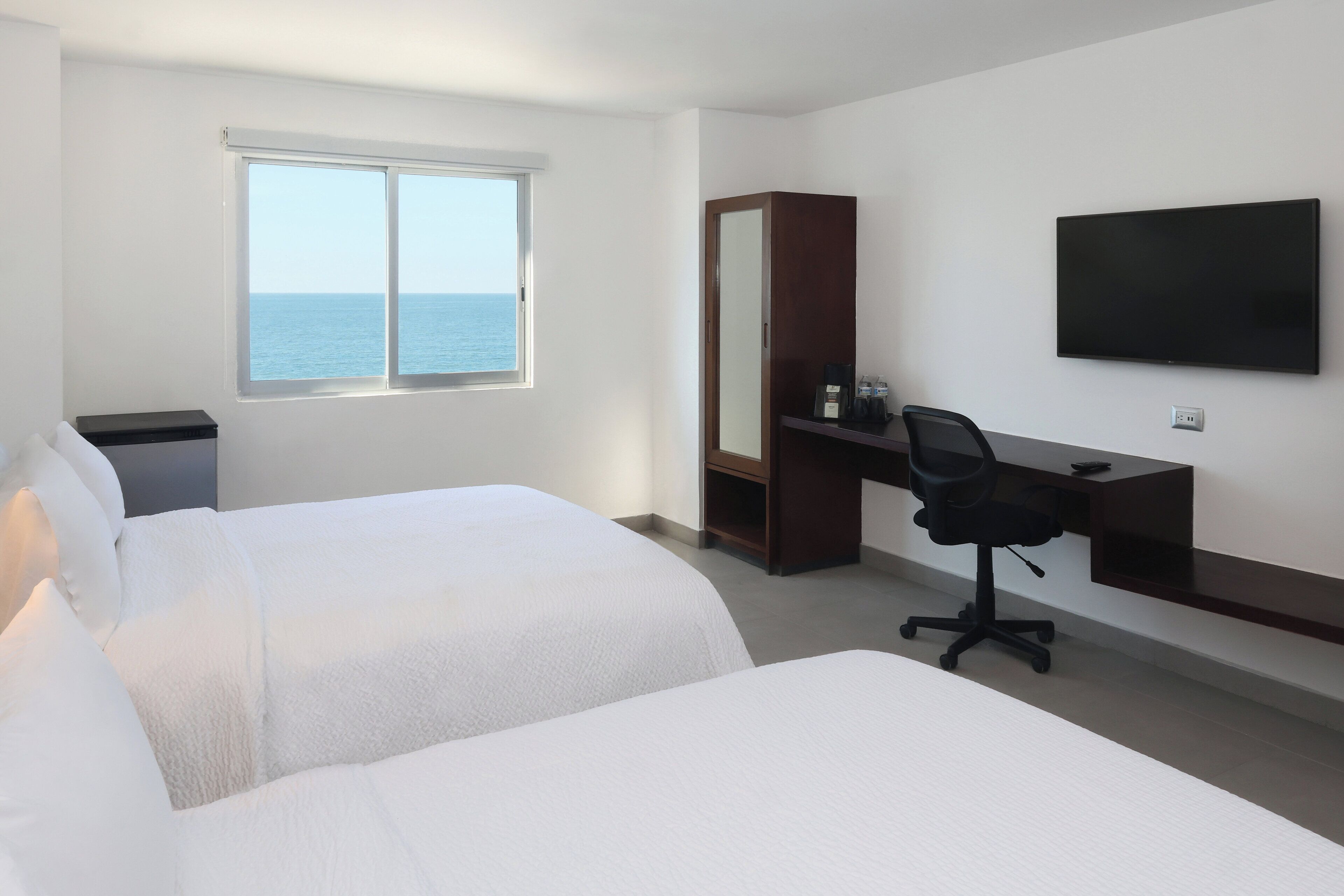 Une chambre d’hôtel avec deux lits, un bureau, une chaise, une télévision et une fenêtre donnant sur la mer.
