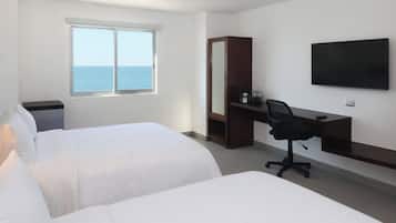 A hotel room with two beds, a desk, a chair, a TV, and a window with a view of the sea.