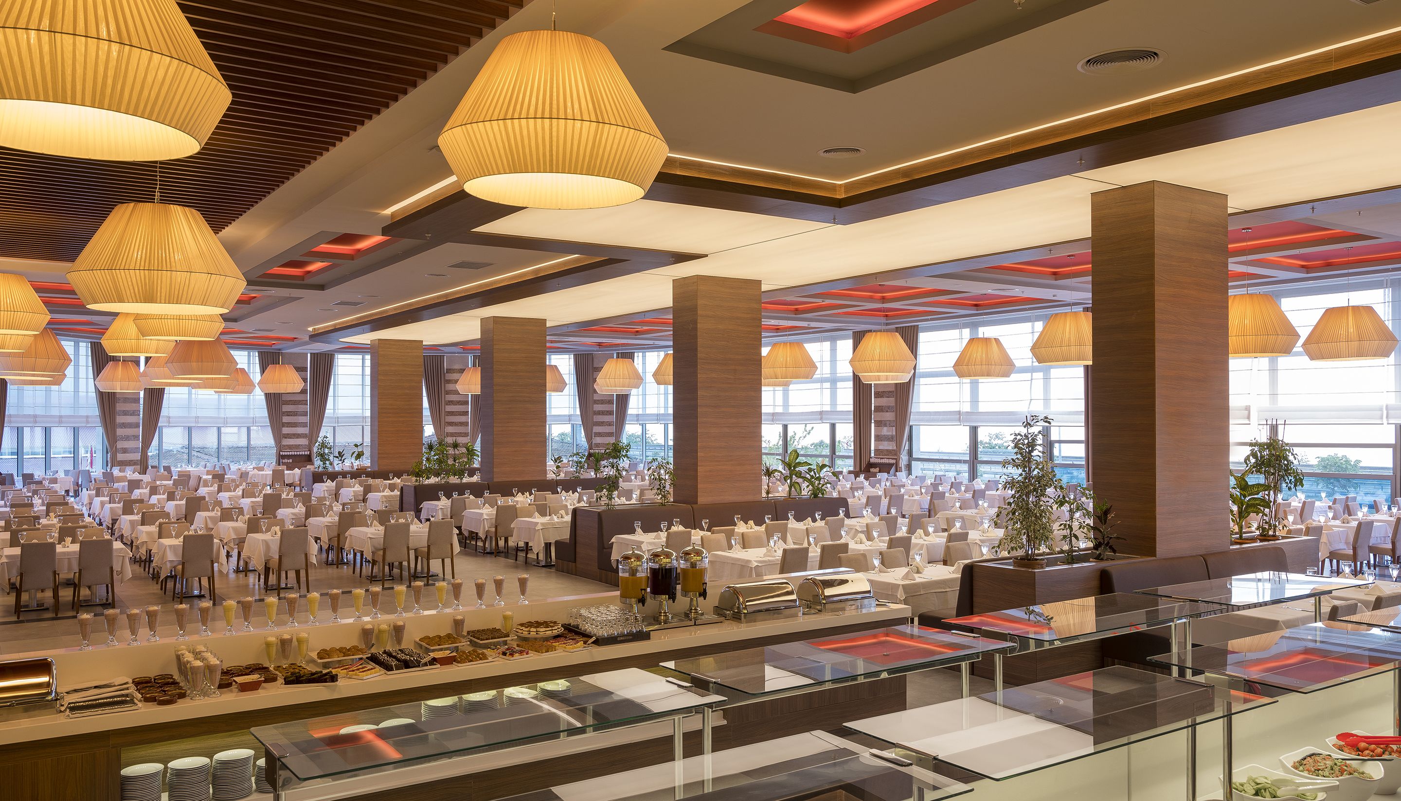 Un amplio salón de banquetes con múltiples mesas dispuestas para comer, grandes lámparas colgantes y una zona de bufé con diversas opciones de comida.