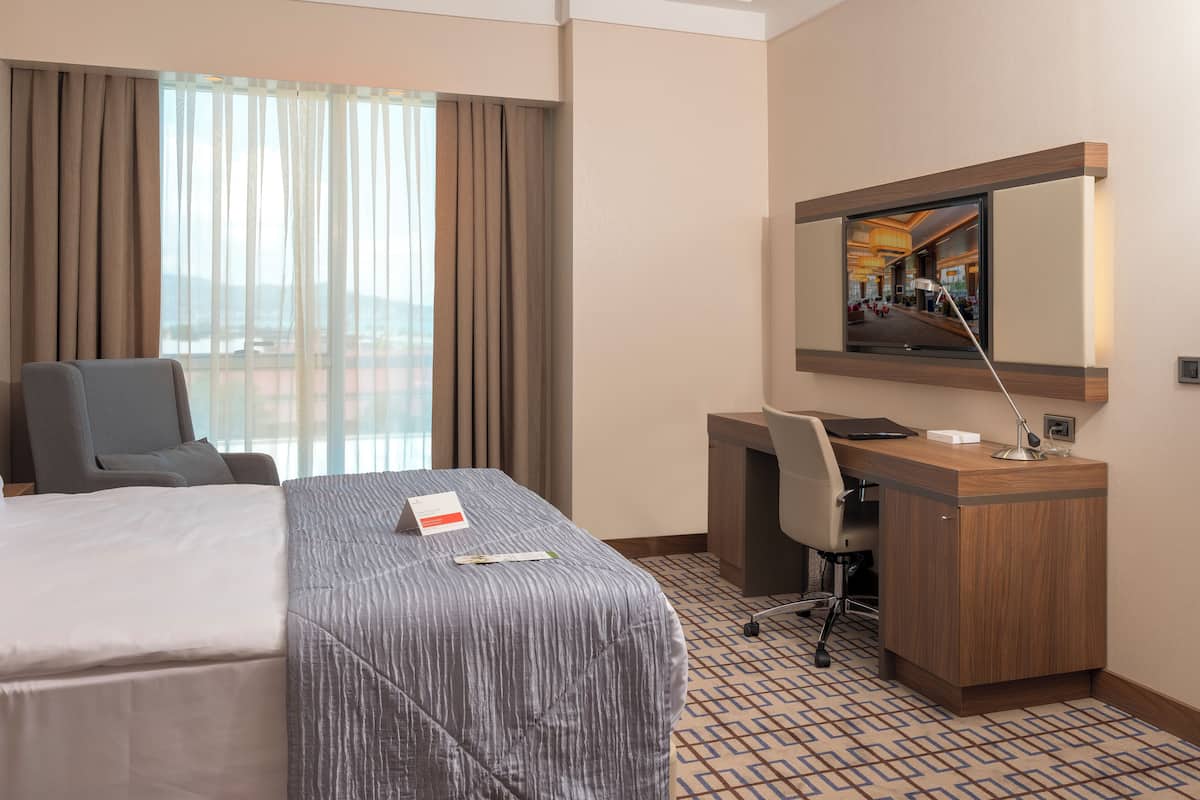 A hotel room with a bed, a desk, a chair, a desk lamp, and a large window with curtains.