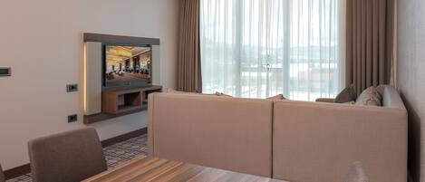 A hotel room with a wooden dining table, beige sofa, and a TV mounted on the wall.