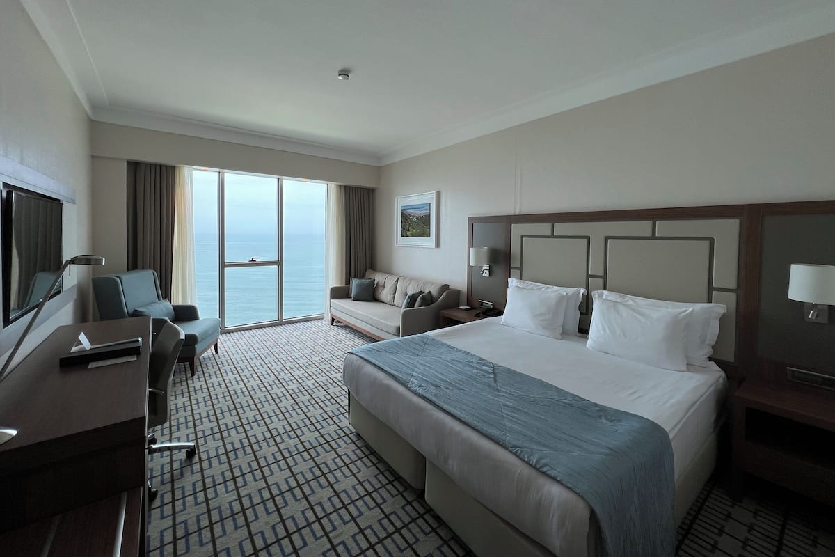 A hotel room with a large bed, a desk, a chair, a TV, and a view of the sea.