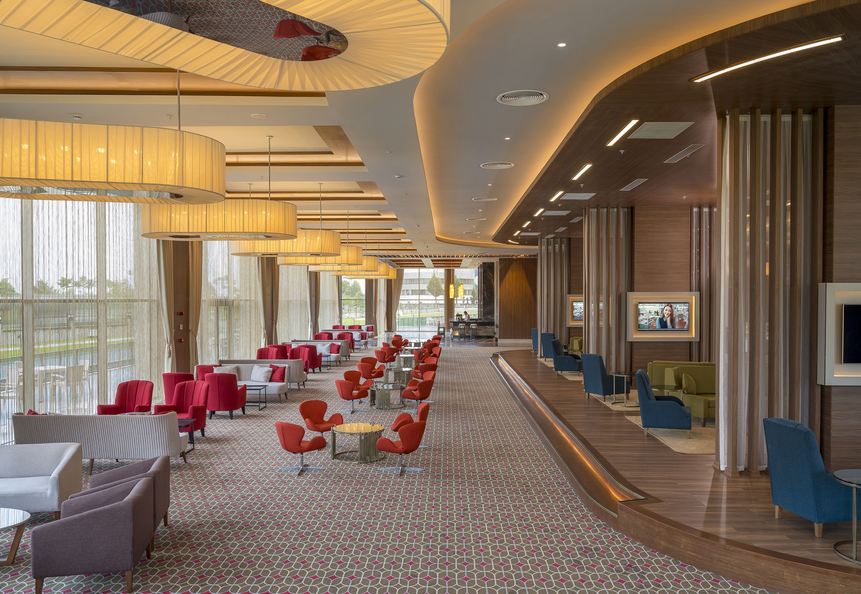 A spacious hotel lobby with a long row of red and blue seating, large windows, and a TV screen.